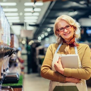 Portrait of happy senior woman holding digital tablet, standing between shelves with food and enjoying her job