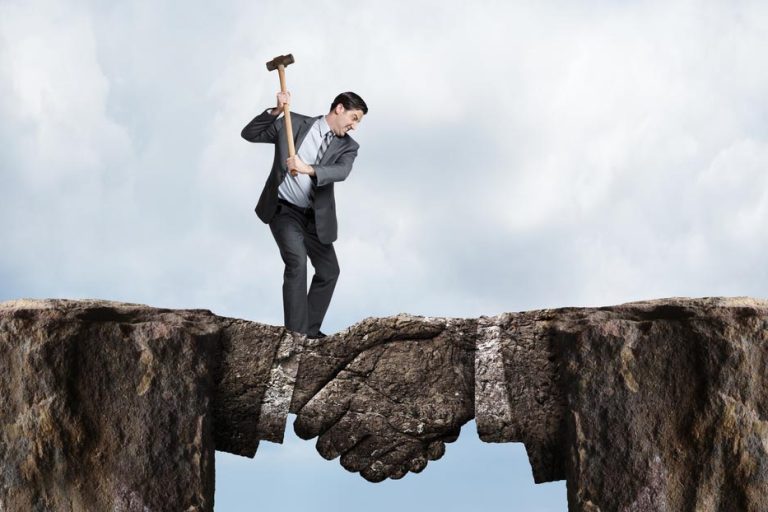 A businessman swings a sledgehammer at a land bridge connecting two cliffs as he tries to chip away at the metaphorical agreement represented by the bridge formed in the shape of a handshake.
