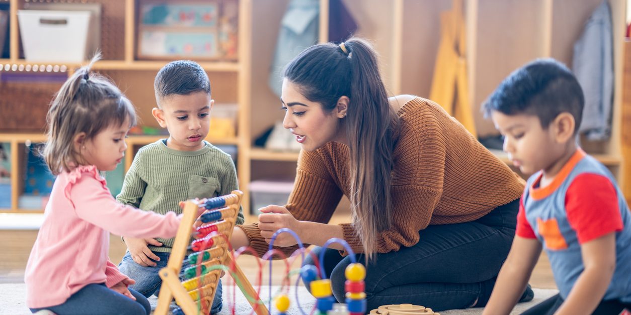 How Your Staff Can Save on Childcare, Health Services A female Kindergarten teacher of Middle Eastern decent, sits on the floor with students as they play with various toys and engage in different activities. They are each dressed casually as they learn through their play.