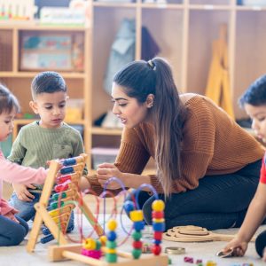 How Your Staff Can Save on Childcare, Health Services A female Kindergarten teacher of Middle Eastern decent, sits on the floor with students as they play with various toys and engage in different activities. They are each dressed casually as they learn through their play.